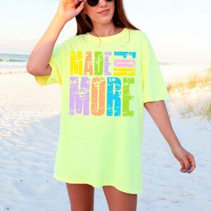 May include: A neon yellow t-shirt with the words "MADE MORE" in a colorful, distressed font. The text is stacked in two lines, with each letter in a different color. The shirt has short sleeves and a relaxed fit.