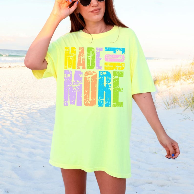 May include: A neon yellow t-shirt with the words "MADE MORE" in a colorful, distressed font. The text is stacked in two lines, with each letter in a different color. The shirt has short sleeves and a relaxed fit.