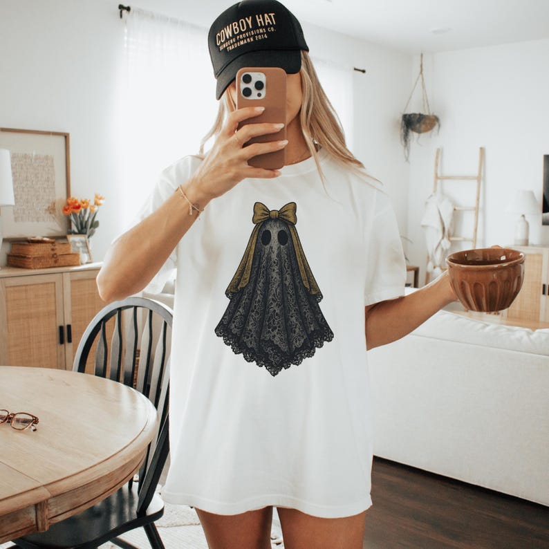 K&ouml;nnte beinhalten: Wei&szlig;es T-Shirt mit einem Geisterdesign. Der Geist hat einen schwarzen, spitzenartigen K&ouml;rper, eine goldene Schleife und einen passenden goldenen Vorhang. Das Design ist auf der Vorderseite des T-Shirts zentriert. Eine schwarze Baseballkappe mit wei&szlig;em Text wird getragen.