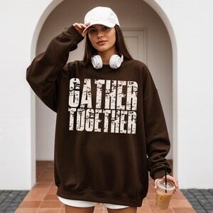May include: A brown sweatshirt with the words "GATHER TOGETHER" in a distressed white font. The sweatshirt is worn with a white baseball cap and white headphones.