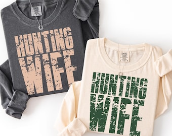 Puede incluir: Se muestran dos camisetas de manga larga, una gris antracita y otra crema. Cada camiseta muestra el texto "HUNTING WIFE" en una fuente desgastada. La camiseta antracita tiene una impresión marrón claro, mientras que la camiseta crema tiene una impresión verde. Ambas camisetas tienen cuello redondo.