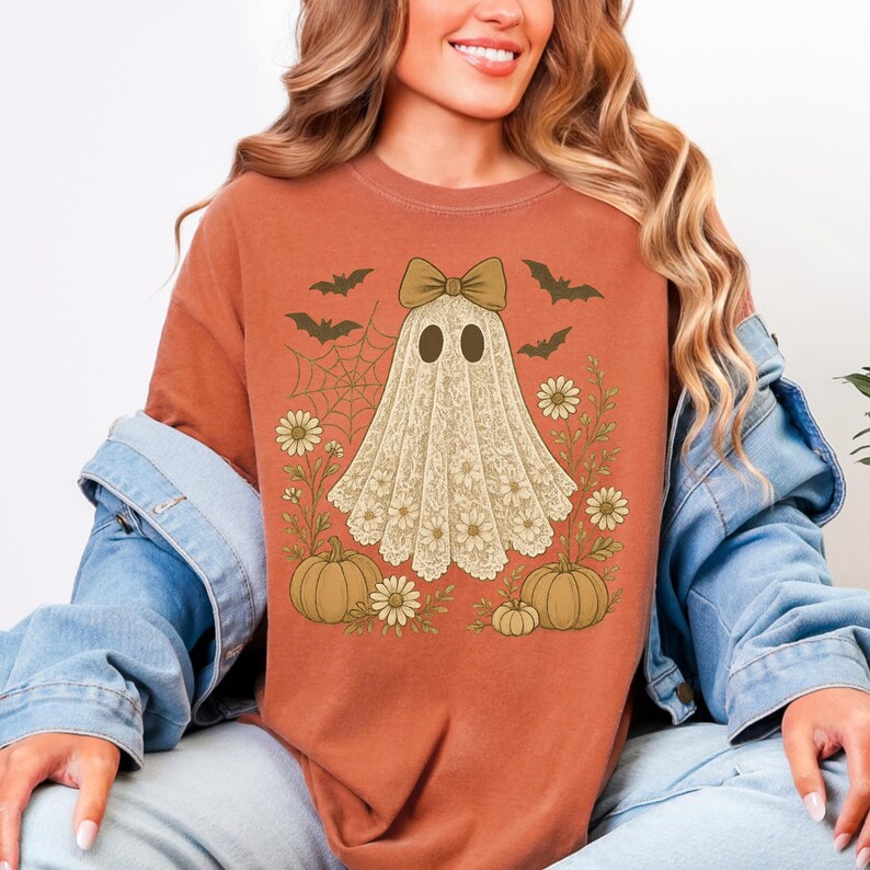 Puede incluir: Camiseta naranja quemado con un dise&ntilde;o de fantasma. El fantasma est&aacute; adornado con un lazo y un patr&oacute;n de encaje floral. El dise&ntilde;o incluye murci&eacute;lagos, una telara&ntilde;a, margaritas y calabazas, creando un tema de Halloween.