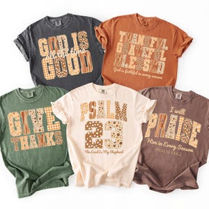 以下が含まれることがあります： さまざまな色の5枚のTシャツ。それぞれにインスピレーションを与えるメッセージが書かれています。テキストは、花柄とチェック柄のパッチワークスタイルです。シャツは、グレー、オレンジ、グリーン、クリーム、ブラウンの色合いです。