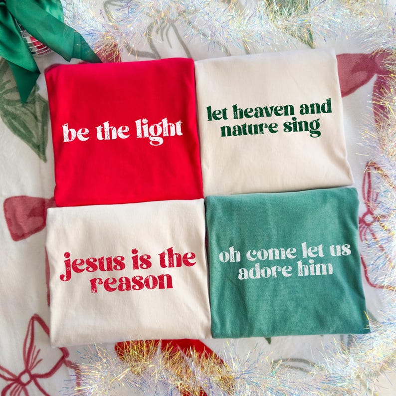 Puede incluir: Cuatro camisetas plegadas en rojo, crema y verde azulado. Cada camiseta presenta un texto diferente en blanco o rojo: "be the light", "let heaven and nature sing", "jesus is the reason" y "oh come let us adore him".