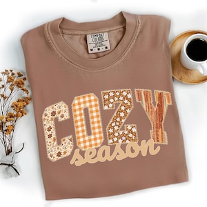 May include: A brown Comfort Colors t-shirt with the words "COZY season" in a patchwork design. The letters have floral, plaid, and striped patterns. A cup of coffee and dried flowers are in the background.