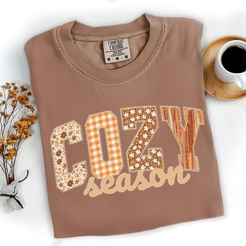May include: A brown Comfort Colors t-shirt with the words "COZY season" in a patchwork design. The letters have floral, plaid, and striped patterns. A cup of coffee and dried flowers are in the background.