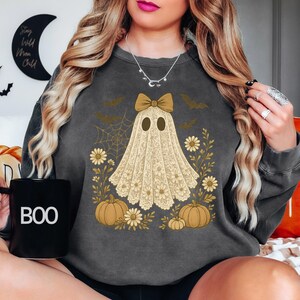 Puede incluir: Sudadera gris oscuro con un dise&ntilde;o de fantasma con lazo, rodeado de calabazas, flores y murci&eacute;lagos. El fantasma es de encaje. Una taza negra con la palabra "BOO" se sostiene en la mano.