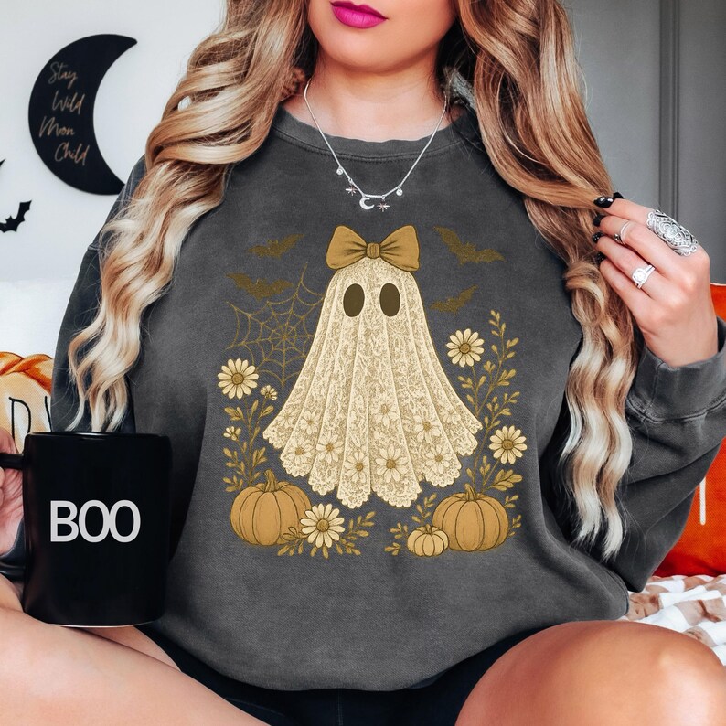 Puede incluir: Sudadera gris oscuro con un dise&ntilde;o de fantasma con lazo, rodeado de calabazas, flores y murci&eacute;lagos. El fantasma es de encaje. Una taza negra con la palabra "BOO" se sostiene en la mano.