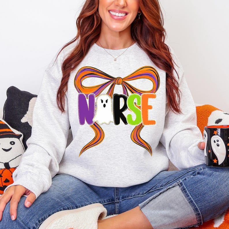 May include: A light gray sweatshirt with the word "NURSE" in purple, white, black, and orange letters, with a ghost graphic. The word is set against a large orange and purple bow. The sweatshirt is part of a Halloween theme.