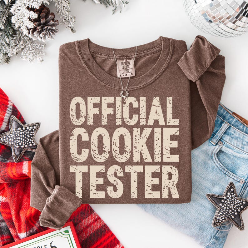 May include: A brown long-sleeve shirt with the text "OFFICIAL COOKIE TESTER" in a distressed, off-white font. The shirt is folded, with a silver necklace visible. A pair of blue jeans and holiday decorations are in the background.