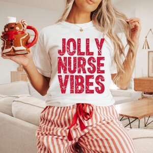May include: White t-shirt with the text "JOLLY NURSE VIBES" in red. The person is wearing red and white striped pajama pants and holding a gingerbread man mug. The mug has a red handle and scarf.