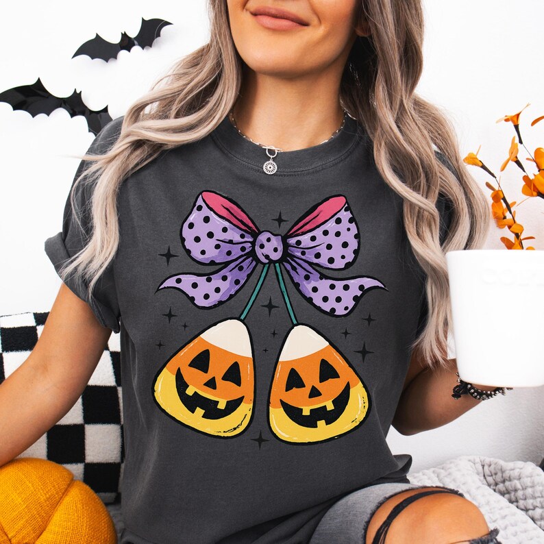 May include: Dark gray t-shirt featuring a Halloween-themed graphic. The design includes two candy corn pieces with jack-o'-lantern faces, connected by green stems to a purple bow with white polka dots. The bow has a pink border.