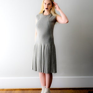 May include: A woman wearing a silver sparkly dress and white ankle boots. She is standing in front of a white wall.