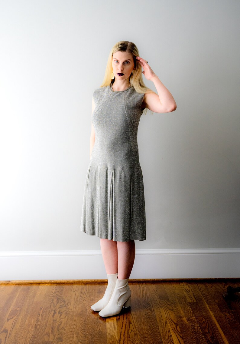 May include: A woman wearing a silver sparkly dress and white ankle boots. She is standing in front of a white wall.