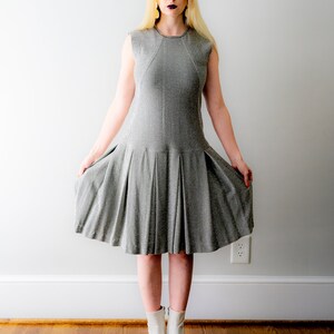 May include: A woman wearing a silver sparkly dress and white ankle boots. She is standing in front of a white wall.