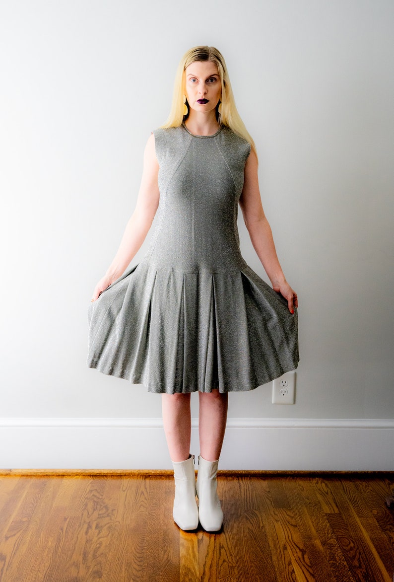 May include: A woman wearing a silver sparkly dress and white ankle boots. She is standing in front of a white wall.