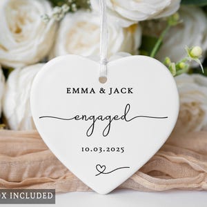 May include: A white heart-shaped ceramic ornament with the words "EMMA & JACK engaged 10.03.2025" in black script. A small heart is at the bottom. The ornament hangs from a white ribbon. White roses and a beige fabric are in the background.