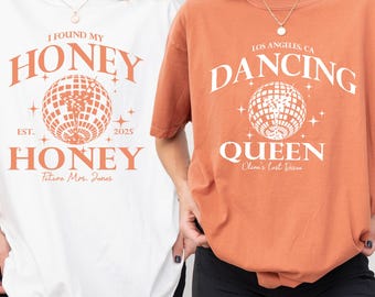 May include: Two oversized t-shirts, one white and one orange, featuring retro-style graphics. The white shirt says "I FOUND MY HONEY" with a disco ball graphic. The orange shirt says "DANCING QUEEN" with a disco ball graphic.