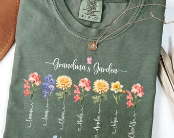 Può includere: T-shirt Comfort Colors verde oliva con la scritta "Grandma's Garden" e i nomi dei propri cari sotto illustrazioni di fiori colorati. Una collana d'oro con un ciondolo poggia sulla maglietta.