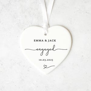May include: A white heart-shaped ornament with a ribbon for hanging. The ornament has the words "EMMA & JACK" above the word "engaged" in cursive, and the date "10.03.2025" below. A small heart is at the bottom.