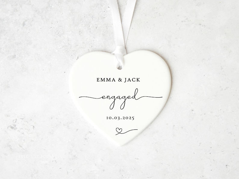 May include: A white heart-shaped ornament with a ribbon for hanging. The ornament has the words "EMMA & JACK" above the word "engaged" in cursive, and the date "10.03.2025" below. A small heart is at the bottom.