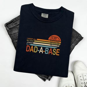 Op de afbeelding: Marineblauw T-shirt met de tekst "I KEEP ALL MY DAD JOKES IN A DAD-A-BASE" in een retro-ontwerp. De afbeelding bevat een diskette en kleurrijke horizontale strepen. Het shirt is opgevouwen op een donkergrijze jeans en witte sneakers.