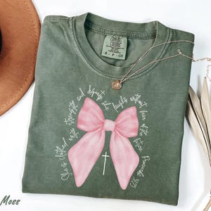 May include: A moss green t-shirt with a large pink bow and a white cross design. The shirt features the text "She is clothed with strength and dignity, she laughs without fear of the future Proverbs 31:25" in a circular pattern. A gold necklace rests on the shirt.