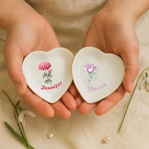 May include: Two heart-shaped, white ceramic trinket dishes with gold rims. Each dish features a floral design and a name in script: one with a pink flower and "Jennifer," the other with a purple flower and "Elizabeth."