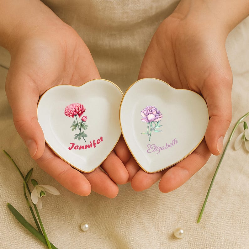 May include: Two heart-shaped, white ceramic trinket dishes with gold rims. Each dish features a floral design and a name in script: one with a pink flower and "Jennifer," the other with a purple flower and "Elizabeth."