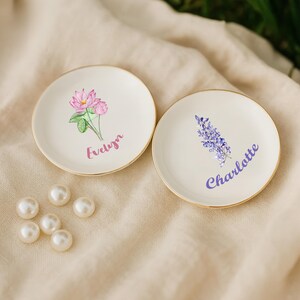 May include: Two small, off-white ceramic plates with gold rims. One plate features a pink floral design with the name "Evelyn" in purple script. The other plate has a purple floral design and the name "Charlotte". Several white pearls are scattered nearby.