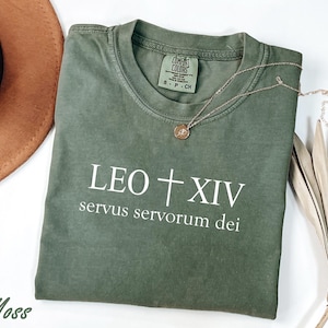 May include: A moss green t-shirt with the text "LEO + XIV servus servorum dei" in white. A gold necklace with a pendant rests on the shirt. The shirt is folded and placed on a white surface.