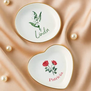 May include: Two decorative trinket dishes. One is round with a floral design and the name "Linda". The other is heart-shaped with a rose design and the name "Patricia". Both have gold rims.
