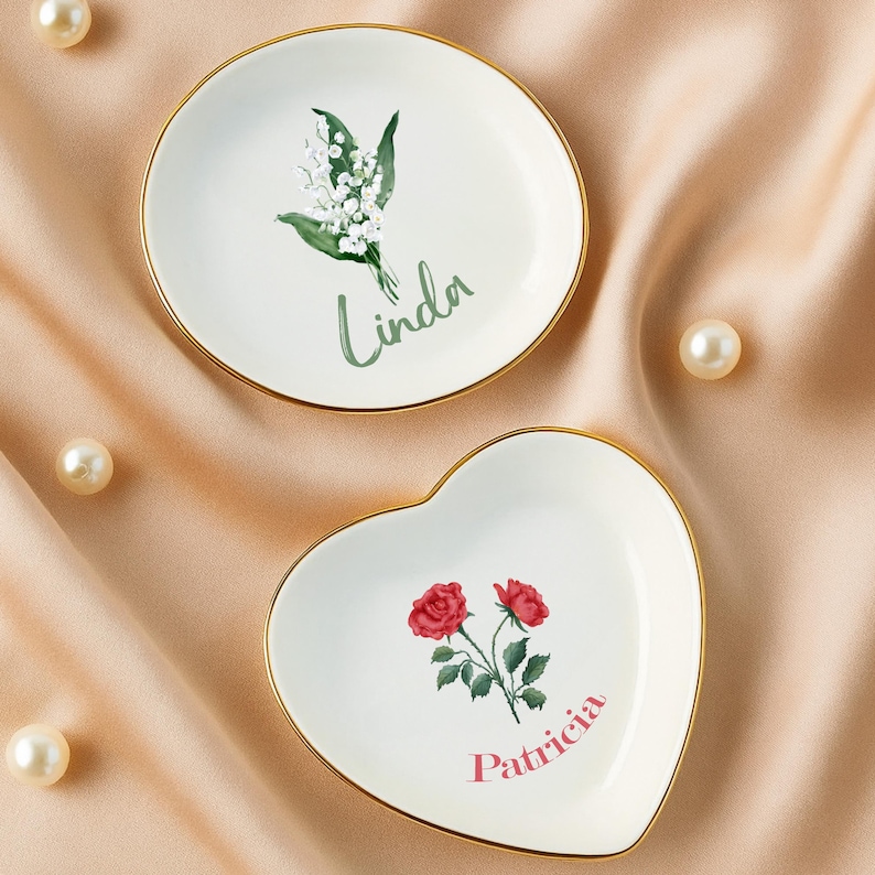 May include: Two decorative trinket dishes. One is round with a floral design and the name "Linda". The other is heart-shaped with a rose design and the name "Patricia". Both have gold rims.