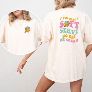 Könnte beinhalten: Cremefarbenes T-Shirt mit einem bunten Eiswaffel-Design auf der Vorder- und Rückseite. Auf der Rückseite des Shirts steht in verschiedenen Farben der Text "IF YOU WANT A SOFT SERVE GO GET ICE CREAM". Auf der Vorderseite befindet sich ein kleines Eiswaffel-Design.