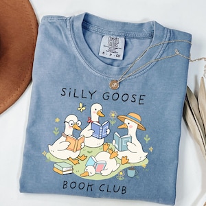 Op de afbeelding: Een blauw Comfort Colors T-shirt met de tekst "SILLY GOOSE BOOK CLUB" en illustraties van stripganzen die boeken lezen. De ganzen zijn omringd door bloemen en een vlinder. Een gouden ketting ligt over het shirt.