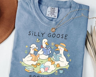 May include: A blue Comfort Colors t-shirt featuring the text "SILLY GOOSE BOOK CLUB" and illustrations of cartoon geese reading books. The geese are surrounded by flowers and a butterfly. A gold necklace is draped across the shirt.