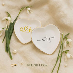 May include: Two heart-shaped white ceramic ring dishes with gold trim. One dish has gold initials and a date, the other has black initials. The dishes are surrounded by flowers, pearls, and wedding rings. Text at the bottom reads "FREE GIFT BOX".