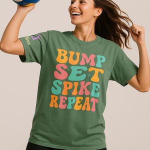 May include: Green t-shirt with the words "BUMP SET SPIKE REPEAT" in colorful, retro-style lettering. A volleyball is held up in the air. The shirt has a crew neck and short sleeves. The person is wearing black shorts.