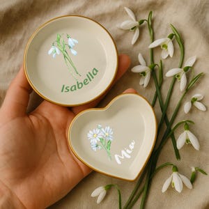 May include: Two small, cream-colored trinket dishes, one round and one heart-shaped, with gold rims. The round dish has a floral design and the name "Isabella" in green. The heart-shaped dish has a floral design and the name "Mia" in white.