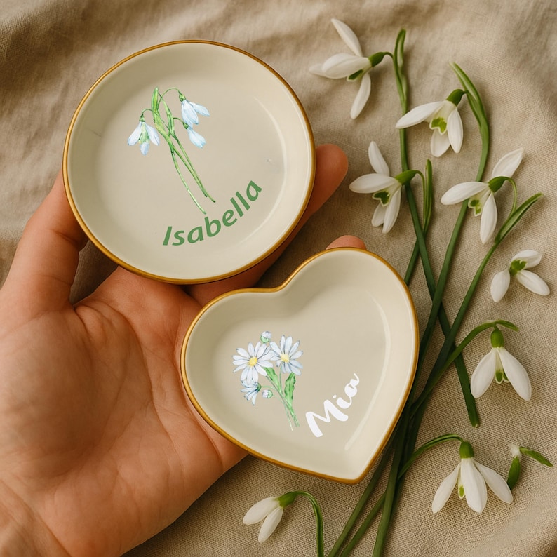 May include: Two small, cream-colored trinket dishes, one round and one heart-shaped, with gold rims. The round dish has a floral design and the name "Isabella" in green. The heart-shaped dish has a floral design and the name "Mia" in white.