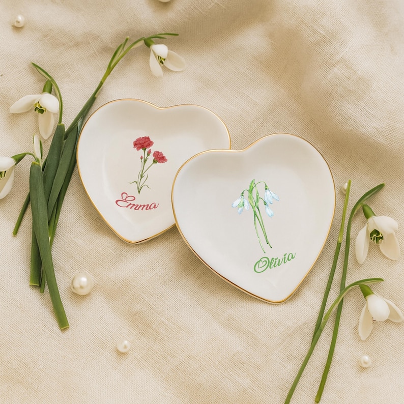May include: Two heart-shaped, white ceramic trinket dishes with gold rims. One features a red flower and the name "Emma" in red script, the other has a blue flower and the name "Olivia" in green script. Snowdrop flowers and pearls surround the dishes.