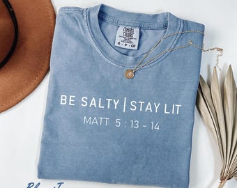 May include: A blue jean colored t-shirt with the text "BE SALTY | STAY LIT" and "MATT 5:13 - 14" printed in white. A gold necklace with a pendant is draped across the shirt. A brown hat is visible in the background.
