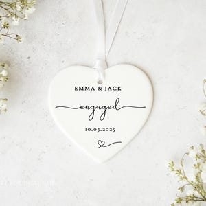 May include: A white heart-shaped ornament with the names "EMMA & JACK" and the word "engaged" in cursive script. The date "10.03.2025" and a heart symbol are also printed on the ornament. A white ribbon is attached for hanging.