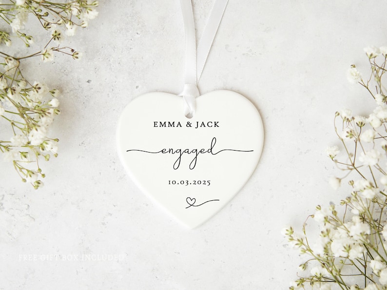 May include: A white heart-shaped ornament with the names "EMMA & JACK" and the word "engaged" in cursive script. The date "10.03.2025" and a heart symbol are also printed on the ornament. A white ribbon is attached for hanging.
