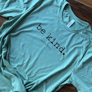 May include: A light teal t-shirt with the words "be kind...or a bitch" printed in black. The shirt is made of soft material and has short sleeves. The shirt is knotted at the bottom.
