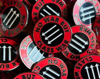 May include: A collection of red and black enamel pins. Each pin features the text "NO WAR BUT CLASS WAR" around a black circle with three white arrows. The pins are round and have a silver border.