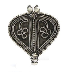 May include: A silver pendant shaped like a heart, featuring intricate floral designs and a beaded border. The pendant has a loop at the top for attaching to a chain.