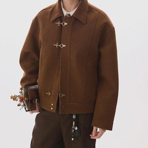 Puede incluir: Chaqueta de lana marrón con cuello con solapas y tres cierres de palanca dorados. La chaqueta tiene un corte entallado y costuras verticales. La persona lleva pantalones marrones y sostiene un pequeño libro marrón con flores.