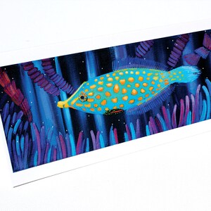 May include: A blue and yellow fish with gold spots swims through a dark blue background with purple and blue seaweed. The fish has a long snout and a small mouth.