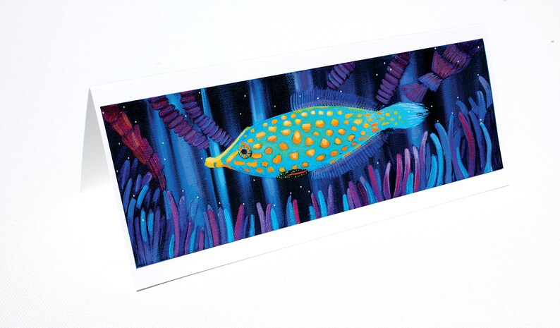 May include: A blue and yellow fish with gold spots swims through a dark blue background with purple and blue seaweed. The fish has a long snout and a small mouth.